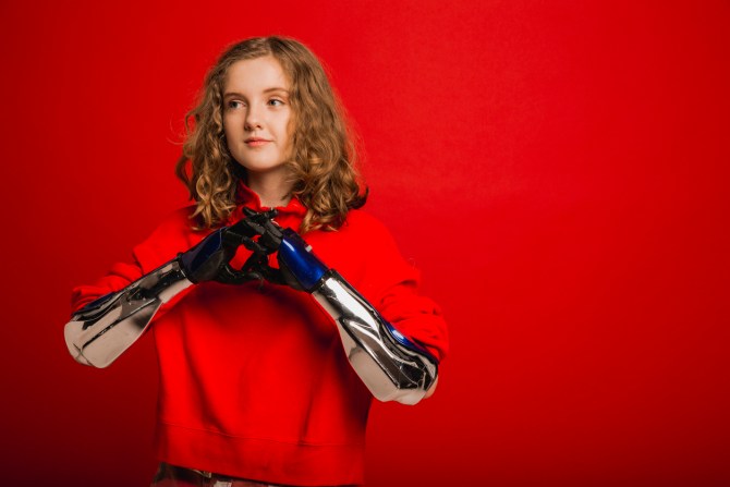 girl with curly blonde hair wearing red hoodie against red background holding prosthetic hands and arms together