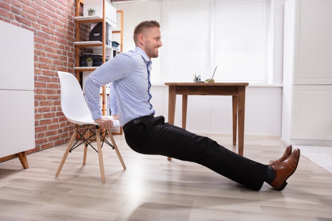 smiling-man-in-business-attire-doing-tricep-dips-in-office-with-white-chair