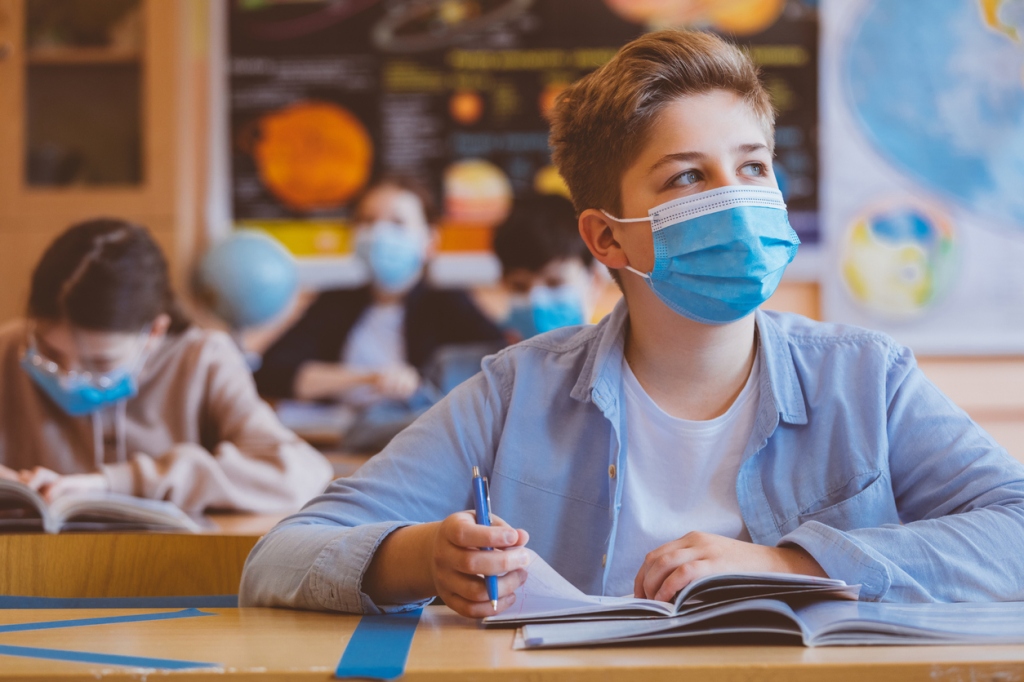 high-school-boy-wearing-blue-surgical-facemask-looking-away-while-sitting-at-desk-during-classroom-learning-period-with-classmates-in-background