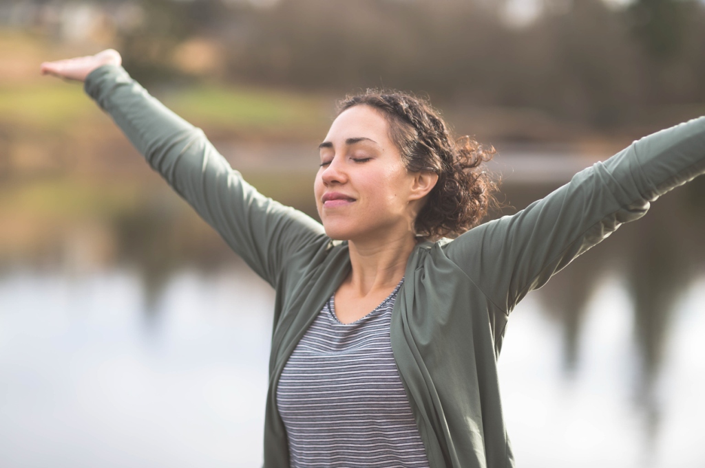 young-woman-wearing-grey-cardigan-raising-arms-while-smiling-outdoors-by-pond