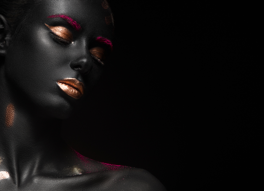 photo of woman with dark skin wearing gold eyeshadow and lipstick looking down while against black background
