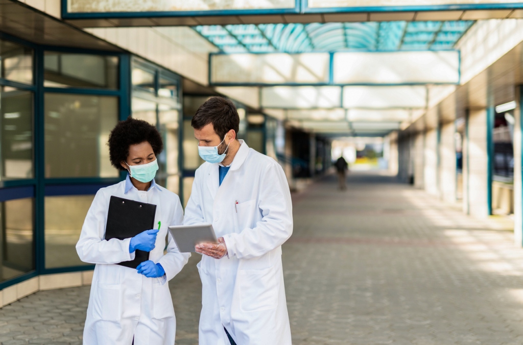 two-doctors-wearing-white-coats-and-face-masks-standing-in-outdoor-corridor-looking-at-tablet