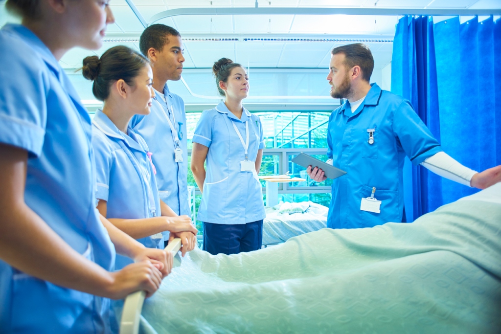 A  senior male staff nurse demonstrates the medical mannequin to a group of medical student nurses . They are all standing around the hospital bed listening to him .