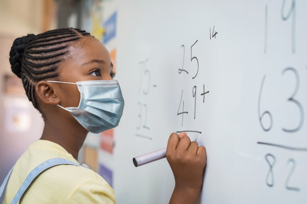 young-girl-wearing-surgical-mask-standing-at-white-board-doing-math-problem-during-classroom-learning